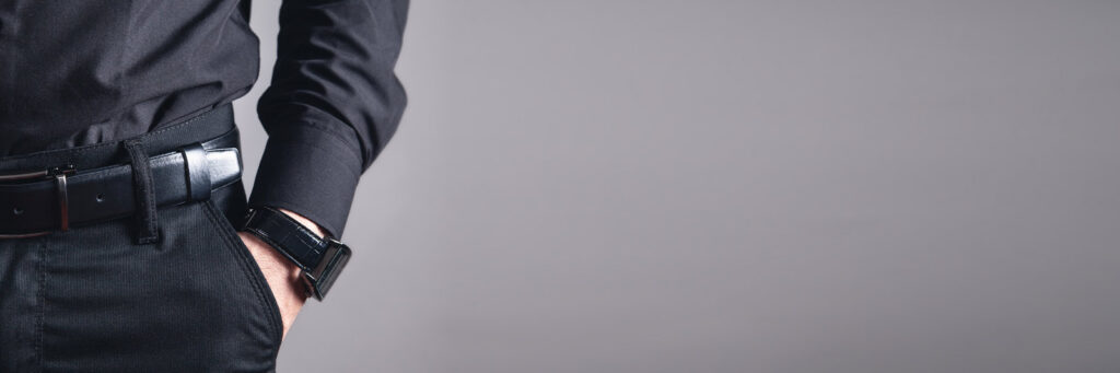 Man wearing a black luxury watch with hand in pocket against a gray background